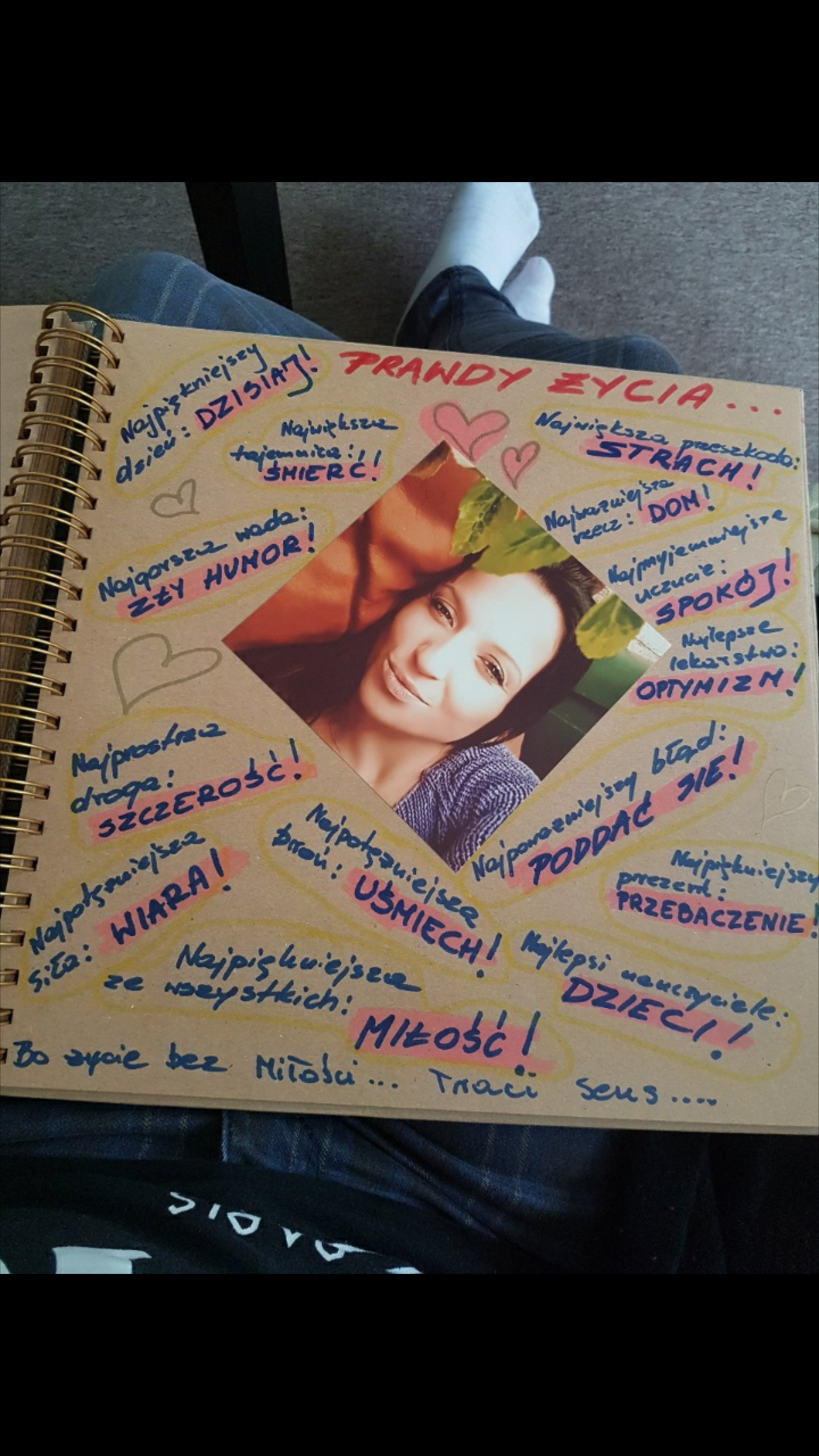 Osobisty notatnik z pozytywnymi afirmacjami i zdjęciem kobiety wklejonym w kształcie rombu, leżący na kolanach osoby w jeansach i białych skarpetkach.