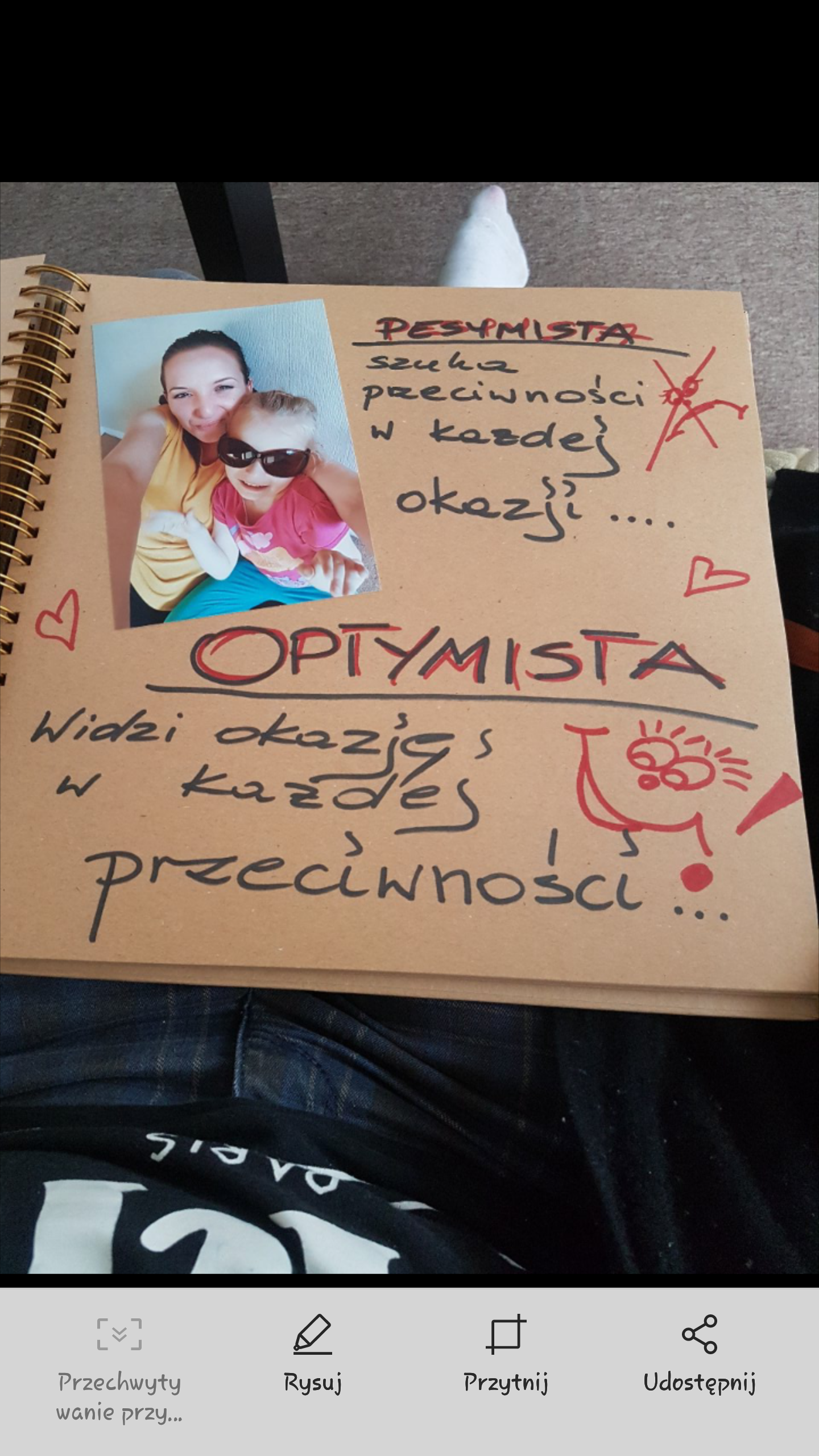 Otwarty notatnik z napisem 'Pesymista szuka przeciwności w każdej okazji' przekreślonym i napisem 'Optymista widzi okazję w każdej przeciwności', obok naklejone zdjęcie kobiety z dzieckiem...
