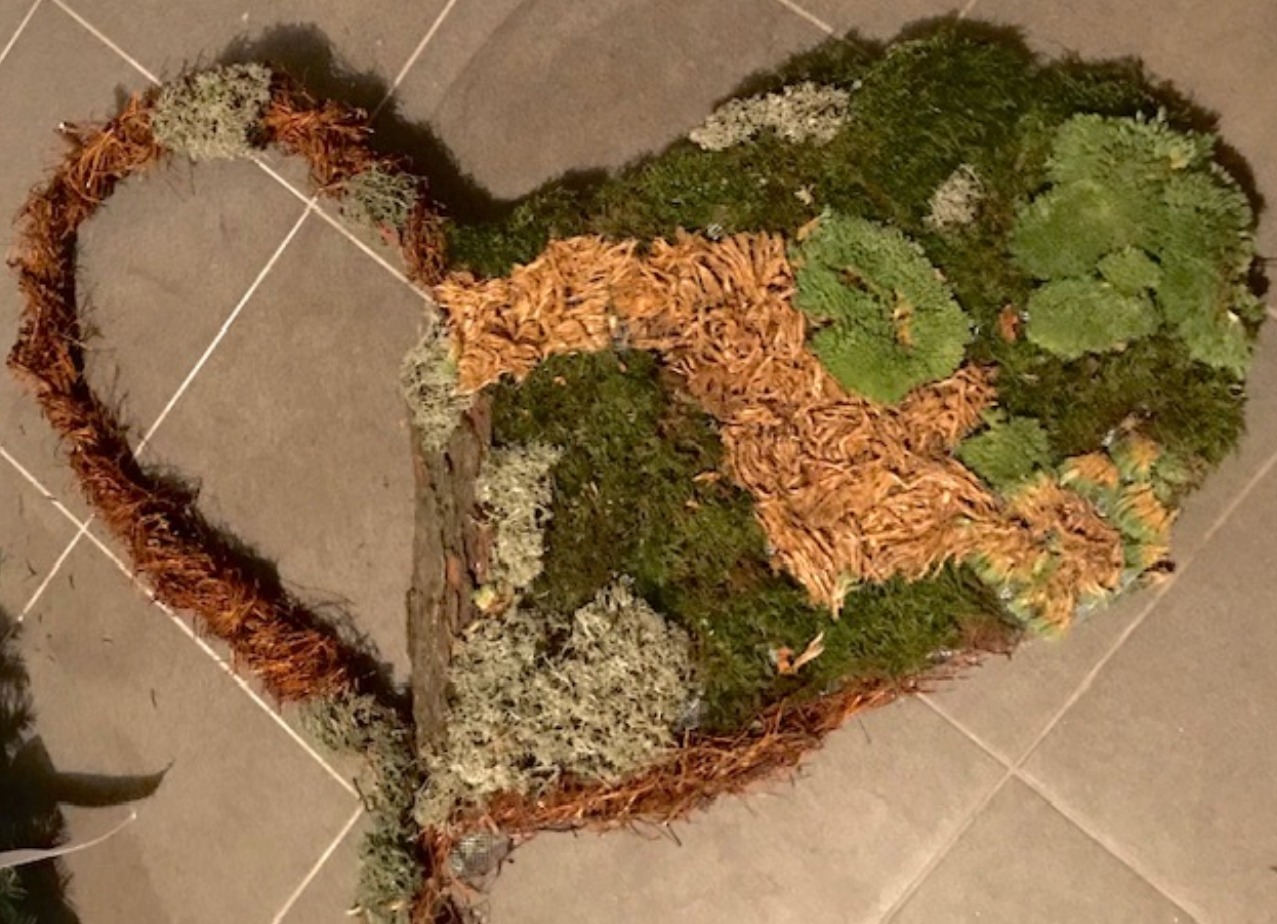 Kompozycja w kształcie serca wykonana z mchu, kory drzewnej i gałązek, leżąca na szarej, kwadratowej płytce podłogowej.