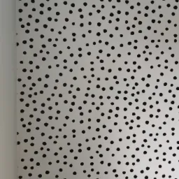 Ściana pokryta białą tapetą w czarne kropki, widoczny fragment listwy przypodłogowej i narożnik ściany, z lekkim cieniem na górze.