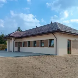 Niski, parterowy budynek z beżową elewacją i ciemną, metalową dachówką, widok z zewnątrz, teren przed budynkiem jeszcze niezagospodarowany.
