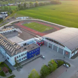 Widok z lotu ptaka na kompleks sportowy z halą widowiskowo-sportową w Bukowcu, boiskiem do piłki nożnej, bieżnią lekkoatletyczną oraz budynkiem szkolnym z niebieskim segmentem.