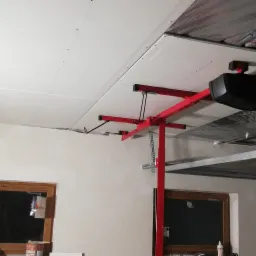 Montaż sufitu z płyty karton gips przy użyciu podnośnika, widoczne łączenia płyt i izolacja termiczna pod stropem. W tle okno i brama garażowa.