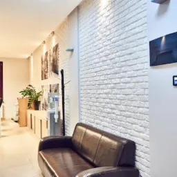 Jasne wnętrze poczekalni z brązową skórzaną sofą, białą ceglaną ścianą, telewizorem i dystrybutorem wody.