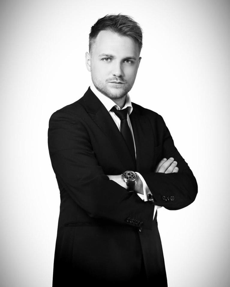 Portret czarno-biały mężczyzny w garniturze z zegarkiem na nadgarstku, skrzyżowane ręce, poważny wyraz twarzy, oświetlenie studyjne.