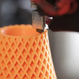 Pomarańczowy, ażurowy model w kształcie ściętego stożka, tworzony przez dyszę drukarki 3D podczas procesu drukowania.