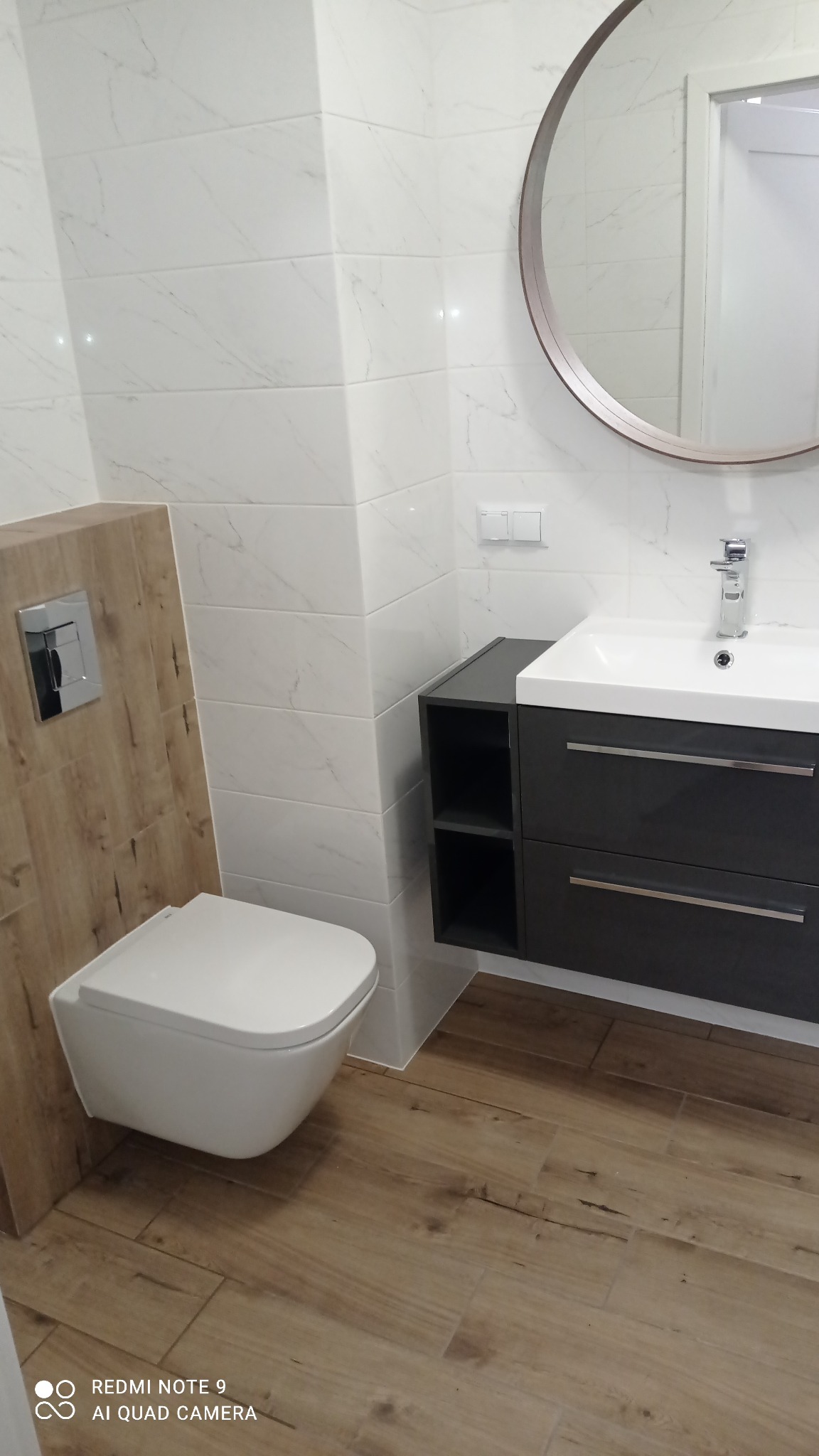 Nowoczesna łazienka z wiszącą toaletą, umywalką z szafką w kolorze grafitowym, okrągłym lustrem i białymi płytkami imitującymi marmur na ścianach oraz drewnopodobnymi płytkami na podłodze.
