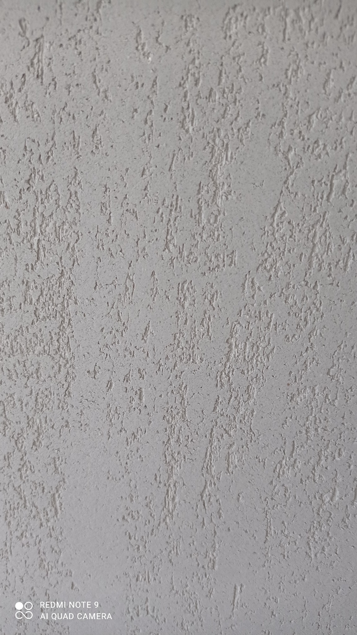 Szara ściana pokryta dekoracyjnym tynkiem strukturalnym z wyraźnym, nieregularnym wzorem wertykalnym.