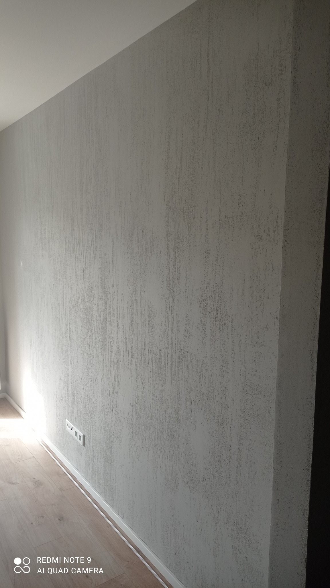 Szara ściana z fakturą, listwa przypodłogowa i gniazdka elektryczne, fragment drewnianej podłogi, widok z dołu.