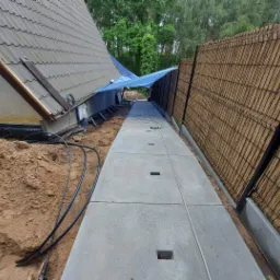 Ułożona betonowa ścieżka z odpływami wzdłuż ogrodzenia z bambusowej maty, widok na fragment domu z zabezpieczeniem folią budowlaną.