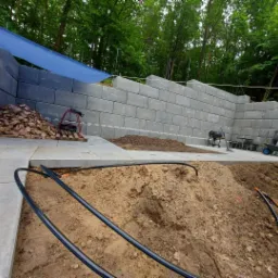 Wznoszenie muru oporowego z betonowych bloków w Gołańcz, widoczny stos materiałów budowlanych i przygotowane podłoże.