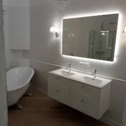 Elegancka łazienka w jasnych kolorach z wolnostojącą wanną na ozdobnych nóżkach, podwójną umywalką z szafką i podświetlanym lustrem.