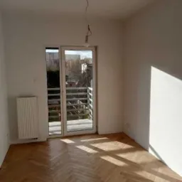 Pusty pokój z parkietem w jodełkę, białymi ścianami, grzejnikiem i balkonem z widokiem na miasto, z wiszącą żarówką bez klosza.