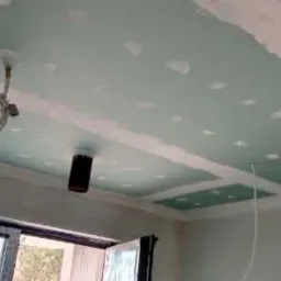 Sufit z płyt gipsowo-kartonowych w trakcie szpachlowania, widoczne łączenia i punkty mocowania, instalacja elektryczna wystająca z sufitu, okno w tle.