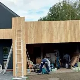 Wznoszenie drewnianej konstrukcji garażu przylegającego do nowoczesnego domu z ciemną elewacją, widoczna drabina i pracownicy w trakcie montażu desek.