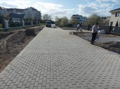 Ułożona szara kostka brukowa na długim podjeździe, widoczni dwaj pracownicy w oddali i budynki mieszkalne w tle.