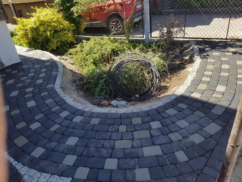 Ułożona ścieżka z kostki brukowej w odcieniach szarości, obrzeże z jasnego kamienia, w tle roślinność i czerwony samochód za ogrodzeniem.