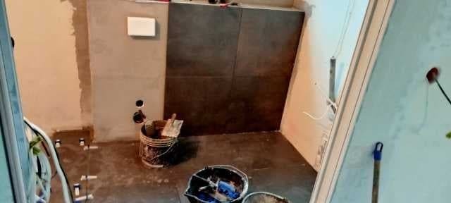 Proces kładzenia ciemnych płytek na ścianie w łazience, widoczne wiadra z zaprawą, szpachelka i krzyżyki dystansowe na podłodze.