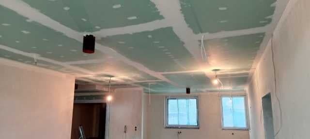 Wnętrze w trakcie wykańczania: sufit z płyt gipsowo-kartonowych, widoczne puszki instalacyjne do oświetlenia i przewody, ściany przygotowane do malowania, okna z niebieską folią ochronną.