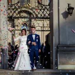 Para młoda wychodzi z kościoła w deszczu konfetti, panna młoda w koronkowej sukni z bukietem, pan młody w granatowym garniturze z muszką, w tle zdobiona brama i uśmiechnięta kobieta z wyrzutnią...