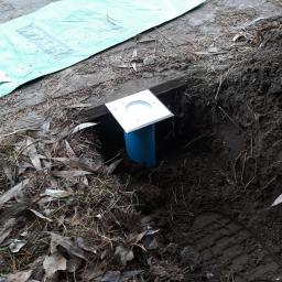 Master Instal - Przygotowanie do montażu oświetlenia zewnętrznego: wykopany otwór z wystającą niebieską rurą i kwadratową, metalową obudową lampy, obok leży zielona folia ochronna na kostce brukowej.