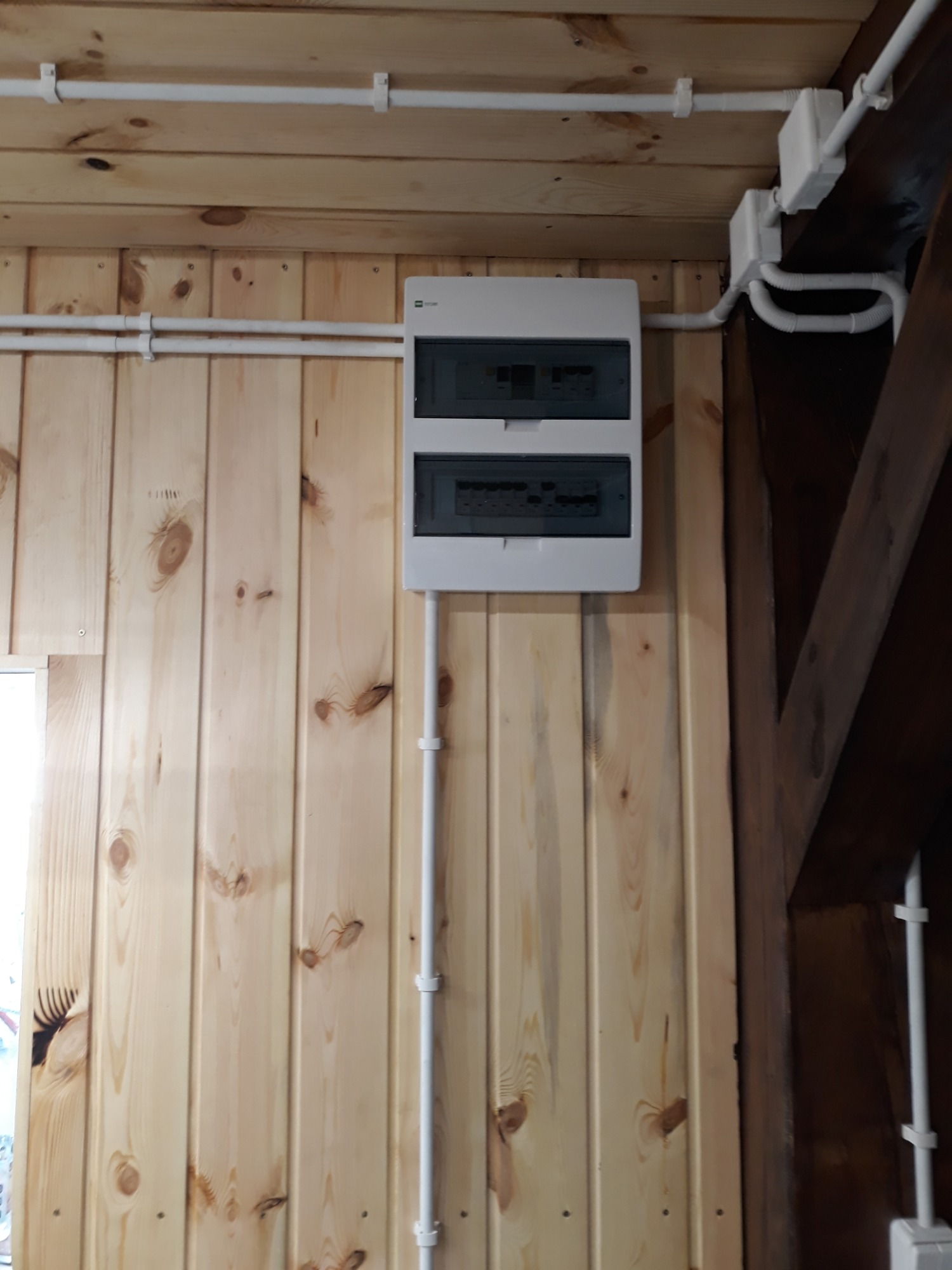 Rozdzielnica elektryczna natynkowa w obudowie z tworzywa, zamontowana na drewnianej ścianie boazeryjnej, z widocznymi rurkami instalacyjnymi w kolorze białym.