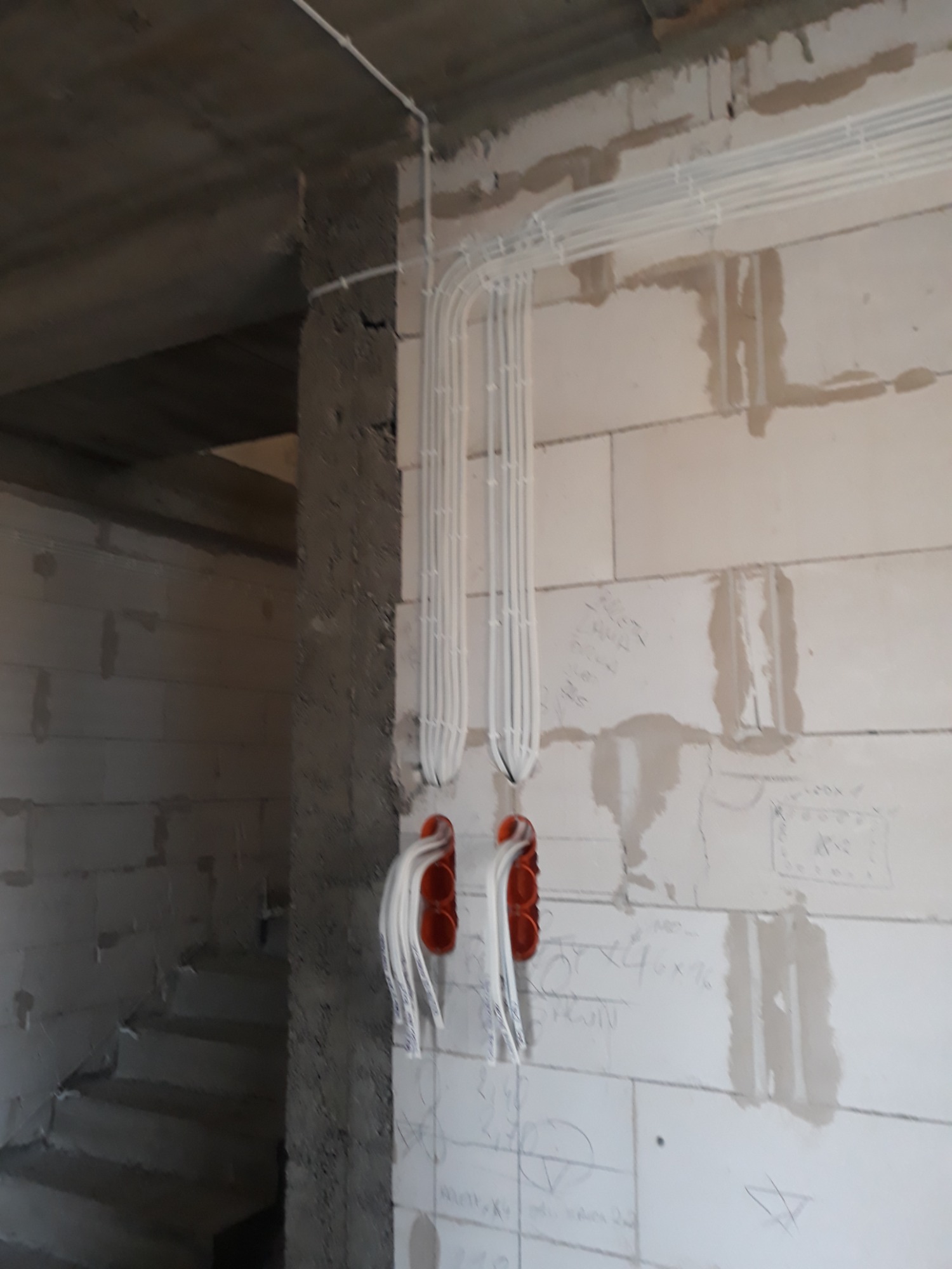 Białe rurki instalacyjne prowadzone po surowej ścianie z betonu komórkowego, widoczne pomarańczowe puszki elektryczne pod tynk, instalacja w trakcie montażu.