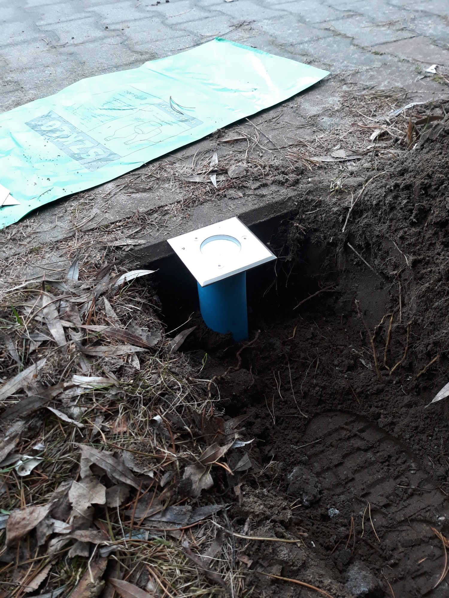 Przygotowanie do montażu oświetlenia zewnętrznego: wykopany otwór z wystającą niebieską rurą i kwadratową, metalową obudową lampy, obok leży zielona folia ochronna na kostce brukowej.