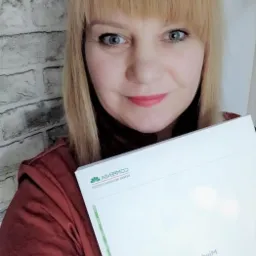 Kobieta z blond włosami trzyma w rękach dokumenty firmy ubezpieczeniowej Compensa, uśmiechając się do kamery na tle ściany z cegły.