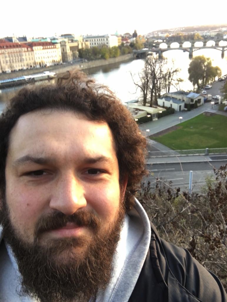 Mężczyzna z brodą robi selfie na tle Wełtawy i mostów w Pradze. Nietypowy kadr z podróży. Fotografia wykonana we Wrocławiu.