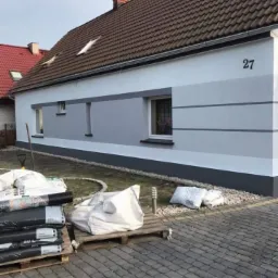 Odnowiona elewacja domu z numerem 27, pomalowana w odcieniach szarości, z workami materiałów budowlanych na paletach przed budynkiem.