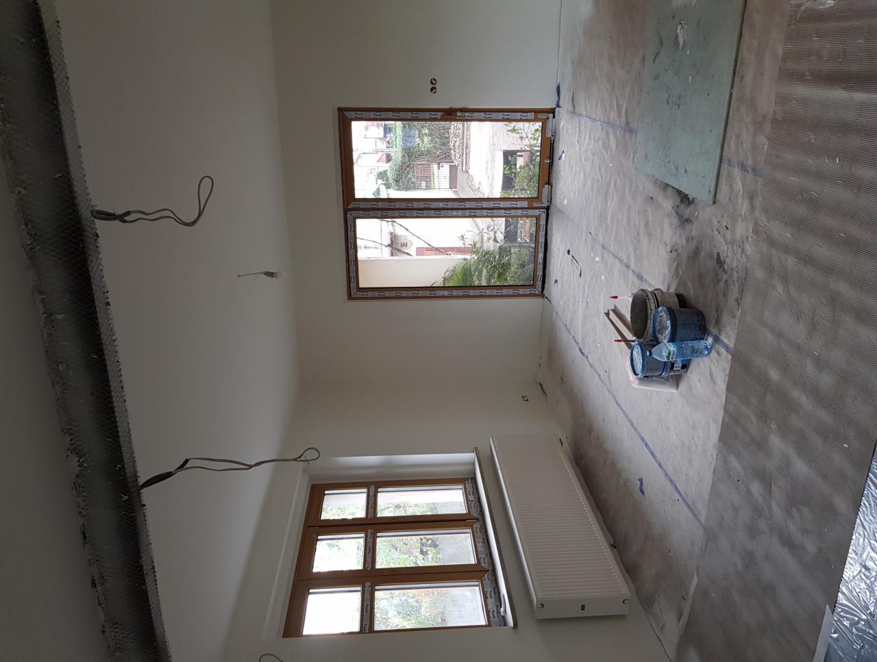 Pokój w trakcie prac wykończeniowych: zabezpieczona podłoga, świeżo pomalowane ściany, widoczne puszki z farbą, grzejnik i dwa okna z drewnianą ramą.