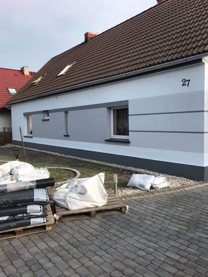 Odnowiona elewacja domu z numerem 27, pomalowana w odcieniach szarości, z workami materiałów budowlanych na paletach przed budynkiem.