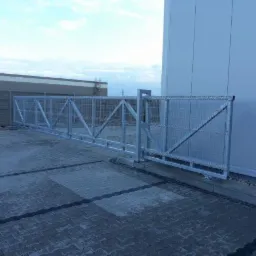 Przesuwna brama wjazdowa z metalowej siatki na szynie, zamontowana przy jasnym budynku przemysłowym i wyłożonym kostką placu.