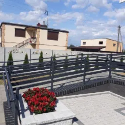 Szara, pozioma balustrada metalowa z poręczą na podjeździe wyłożonym szarą kostką, z widocznymi czerwonymi kwiatami w donicy, w tle dom z betonowym ogrodzeniem panelowym.