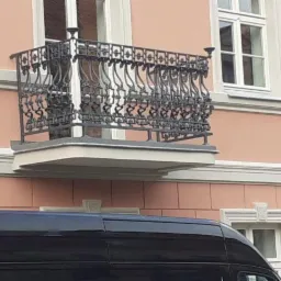 Elegancki balkon z czarną, kutą balustradą na tle różowej fasady budynku z białymi oknami, częściowo zasłonięty przez zaparkowany czarny samochód.