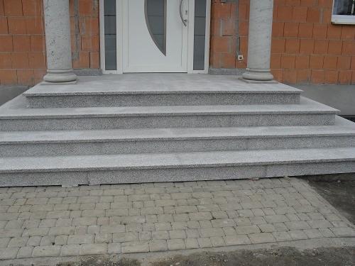 Granitowe schody wejściowe z kolumnami, prowadzące do białych drzwi, z brukowanym chodnikiem przed nimi.