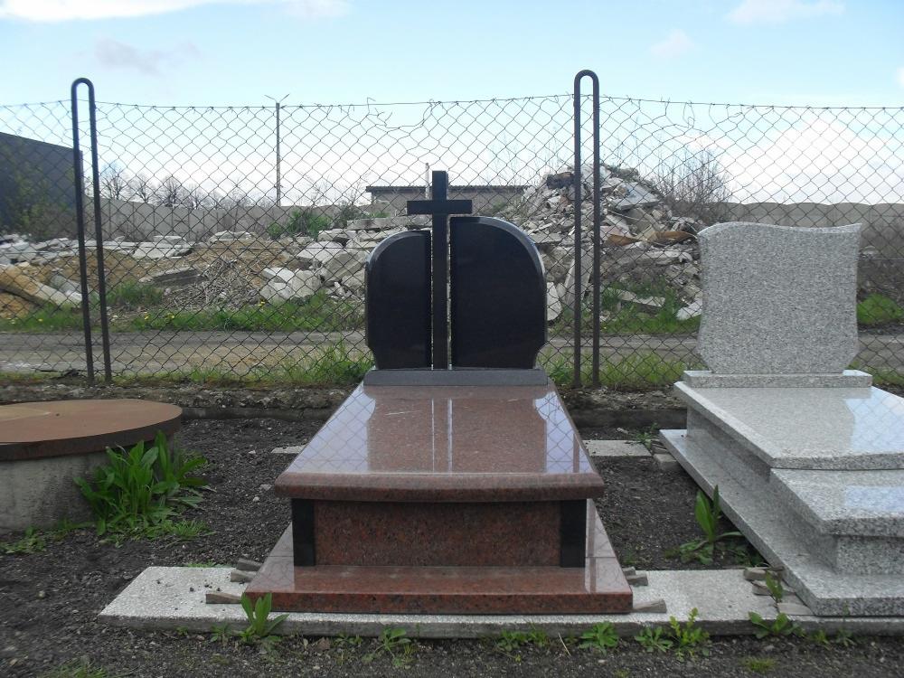 Nowoczesny, polerowany nagrobek z ciemnego granitu z krzyżem, obok jasnoszary nagrobek z delikatnym wzorem, na tle metalowej siatki i sterty gruzu.