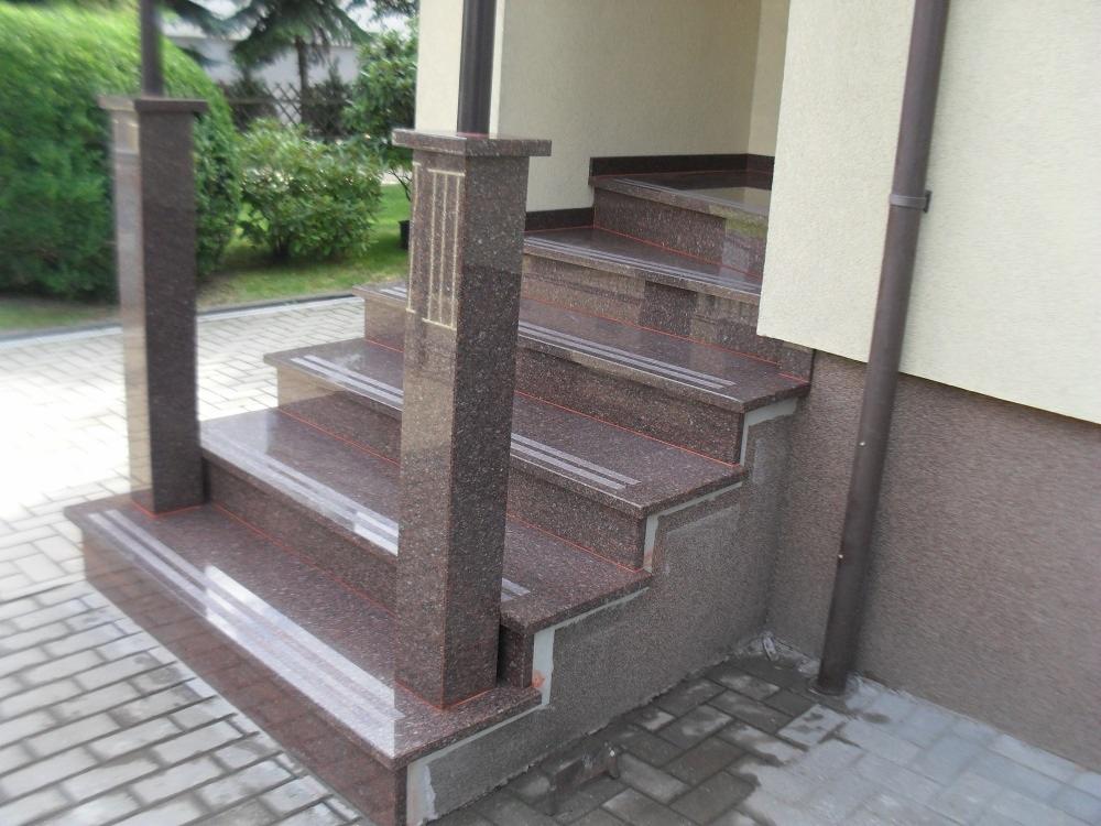 Zewnętrzne schody z brązowego kamienia z dwoma filarami po bokach, wykończone jasnymi paskami antypoślizgowymi, prowadzące do wejścia budynku.