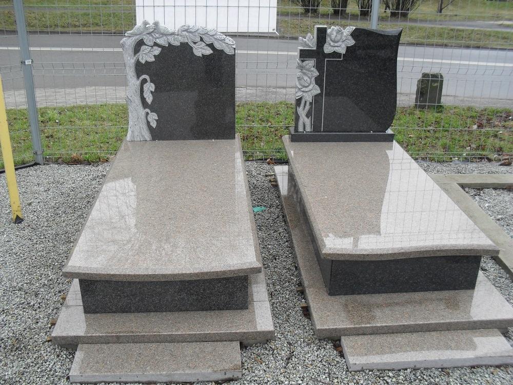 Dwa gotowe nagrobki z ciemnego i jasnego kamienia, jeden z motywem drzewa, drugi z krzyżem i różami, ustawione na żwirowym podłożu.