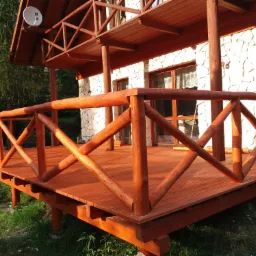 Drewniany taras z balustradą z okrąglaków, przylegający do domu z elewacją z kamienia, z widoczną anteną satelitarną na dachu.