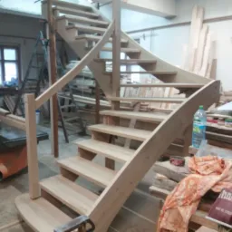 Wewnętrzne, drewniane schody kręcone w trakcie montażu, surowe drewno, widoczne narzędzia i materiały w warsztacie, osoba w tle przy oknie.