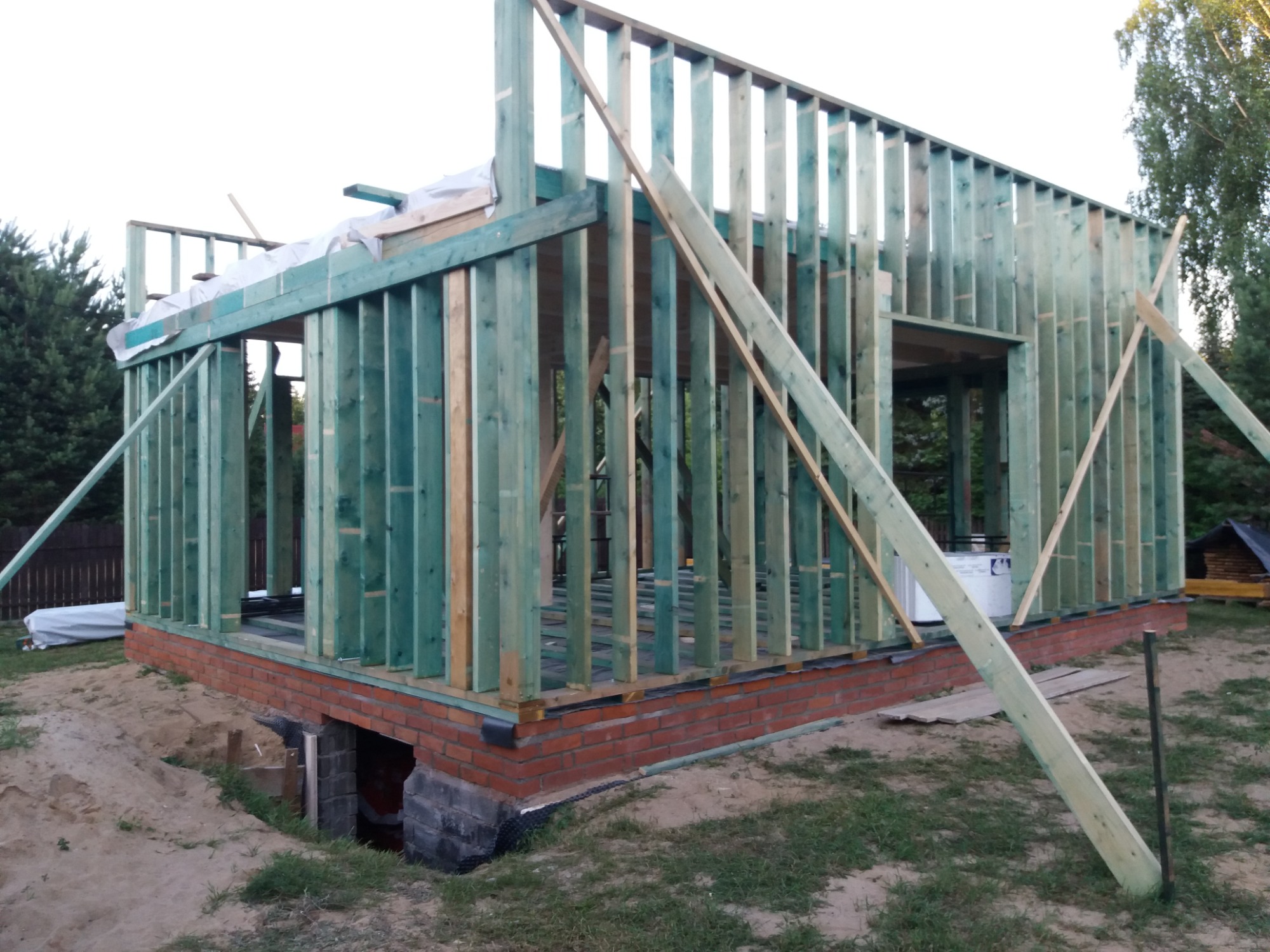 Wznoszenie szkieletu domu: widok na konstrukcję z belek impregnowanych na zielono, osadzoną na fundamencie z cegły, z widocznym wejściem do piwnicy i podporami z desek.