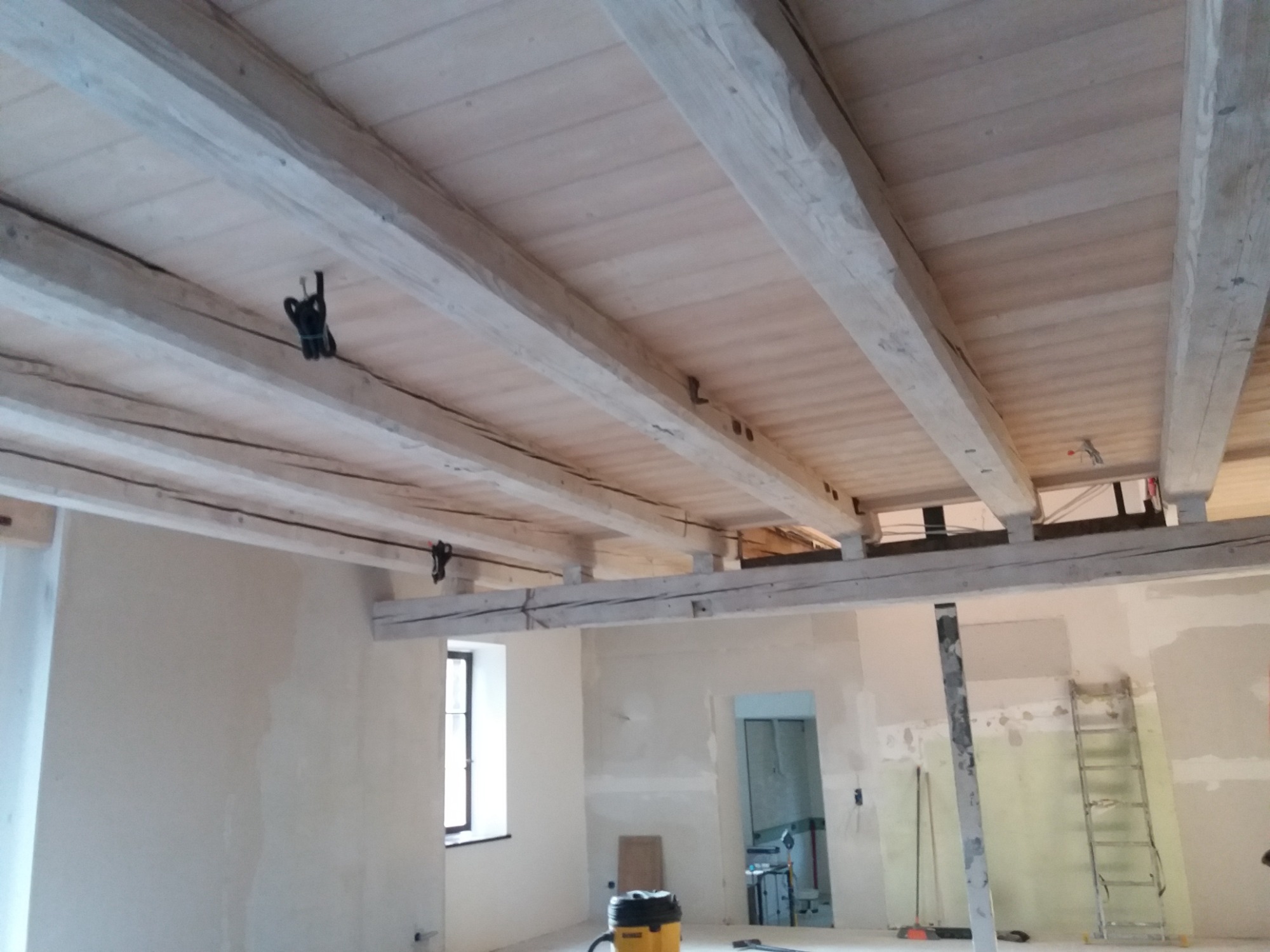 Wnętrze w trakcie remontu z widoczną drewnianą konstrukcją stropu, surowymi ścianami i oknem. Na podłodze widoczny odkurzacz przemysłowy i drabina.