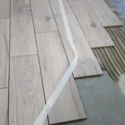 Układanie podłogi z płytek imitujących drewno, widoczne krzyżyki dystansowe i taśma malarska wyznaczająca linię, klej na podłożu.