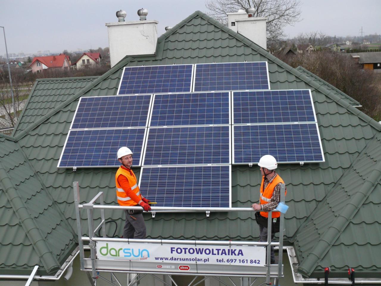 Montaż paneli słonecznych na dachu z zielonej blachodachówki przez dwóch pracowników w kaskach i pomarańczowych kamizelkach, stojących na rusztowaniu z logo firmy Darsun Solar.
