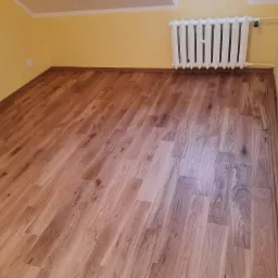 Pokój z nowo ułożonymi panelami podłogowymi o jasnym odcieniu drewna, widoczny biały grzejnik na tle żółtej ściany oraz fragment listwy przypodłogowej.