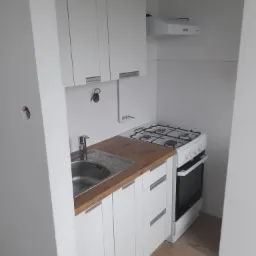 Nowoczesna, biała kuchnia z drewnianym blatem, widok na zlew, kuchenkę gazową z piekarnikiem i biały okap, na ścianie białe płytki, na podłodze panele imitujące drewno, widoczne niedokończone...