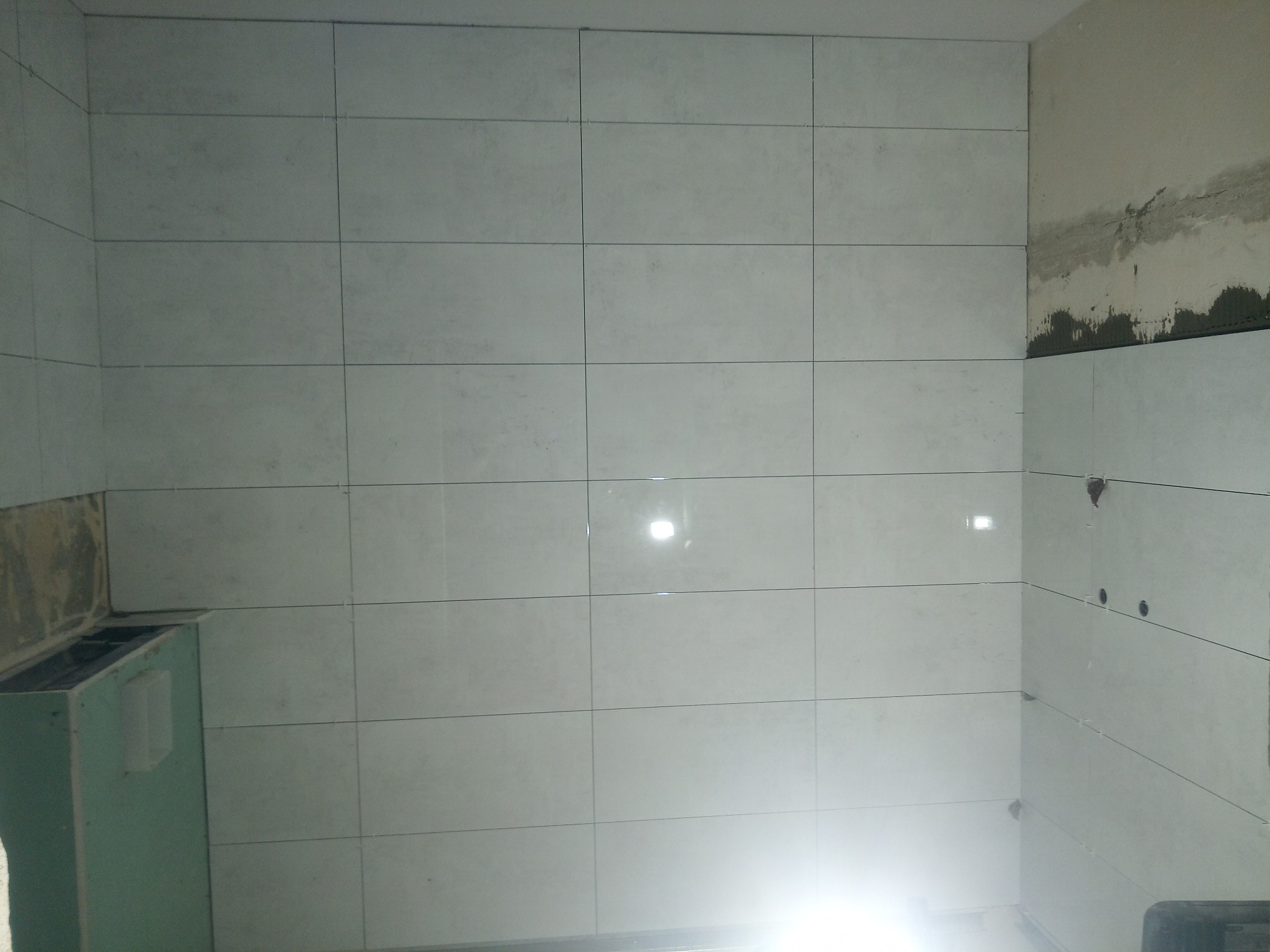 Ściana w trakcie wykańczania z jasnymi, kwadratowymi płytkami ceramicznymi, widoczne nierówności i pozostałości zaprawy na sąsiedniej ścianie, niedokończona zabudowa.