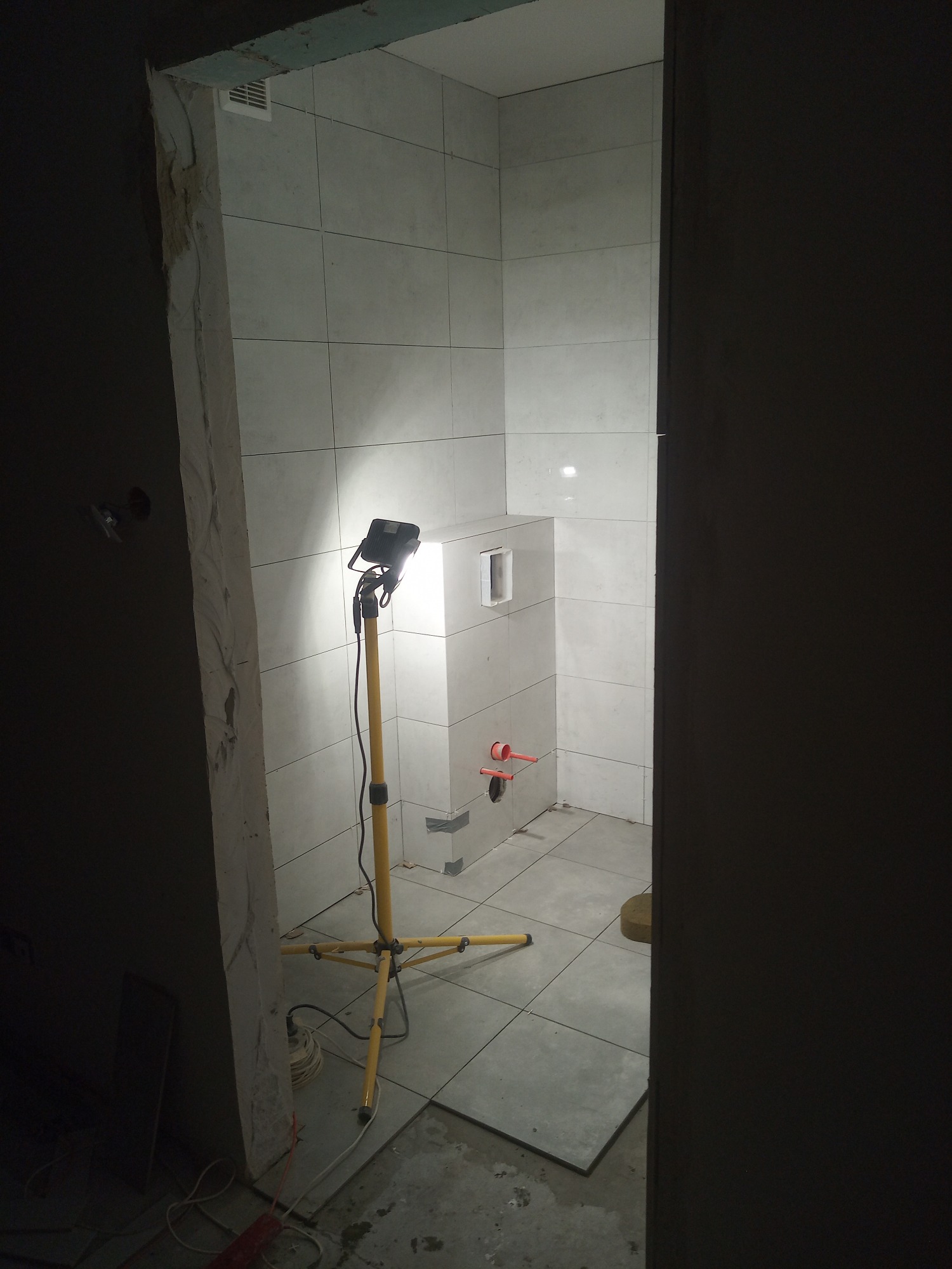 Remontowana łazienka z białymi płytkami, częściowo wykończona, oświetlona lampą budowlaną na trójnogu, widoczne elementy instalacji wodnej i pozostałości po pracach.